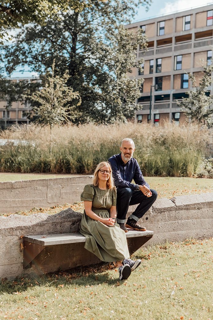 A woman with a green dress and a man with a navy shirt sat outside on a bench with long grass and trees in the background